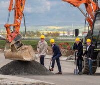 Mehrere Männer im Anzug und Schutzhelm bei einem symbolischen Spatenstich neben einem Bagger.