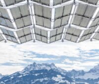 Blick von unten auf die aufgefalteten Photovoltaikmodule eines Solarfaltdachs vor schneebedeckten Bergen.