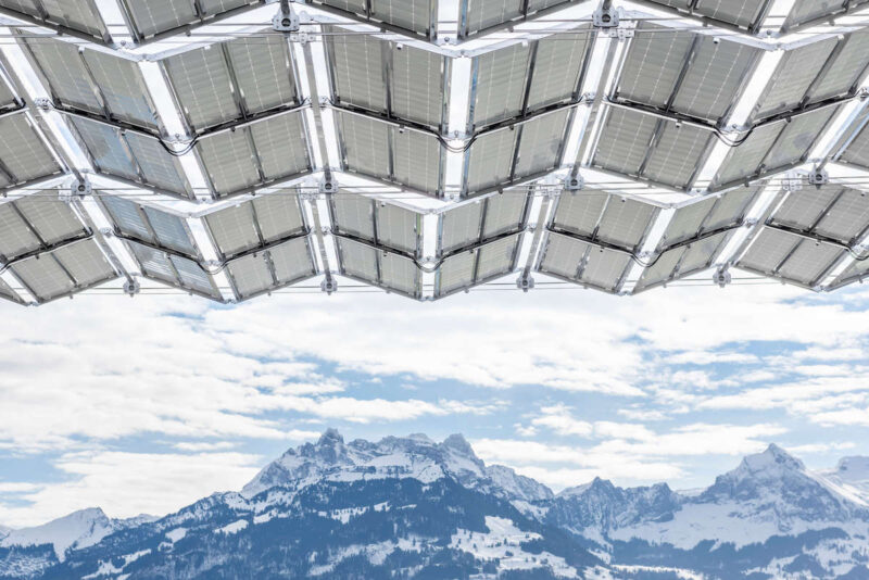 Blick von unten auf die aufgefalteten Photovoltaikmodule eines Solarfaltdachs vor schneebedeckten Bergen.