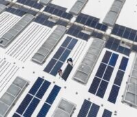Aus der Vogelperspektive ein Blick auf ein Industrie-Leichtdach. Hier montieren Arbeiter gerade die PV-Module