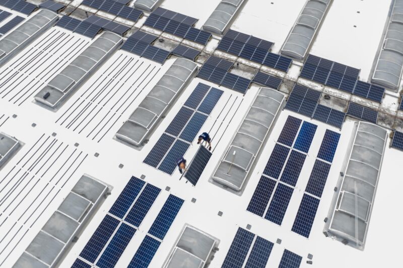 Aus der Vogelperspektive ein Blick auf ein Industrie-Leichtdach. Hier montieren Arbeiter gerade die PV-Module