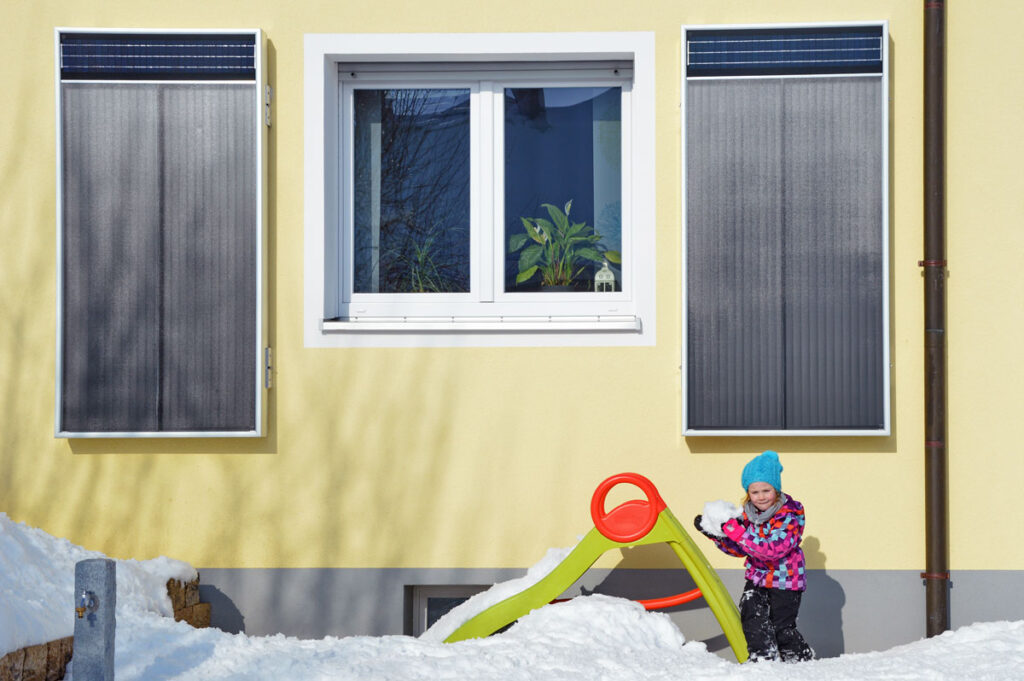 Solar-Luftkollektoren an einer gelben Hauswand montiert. Davor spielt ein Kind im Schnee.