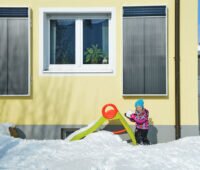 zwei Solar-Luftkollektoren an der Fassade eines Wohnhauses mit spielendem Kind um Schnee