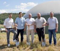 5 Personen mit Spaten vor einem Biogasbehälter.