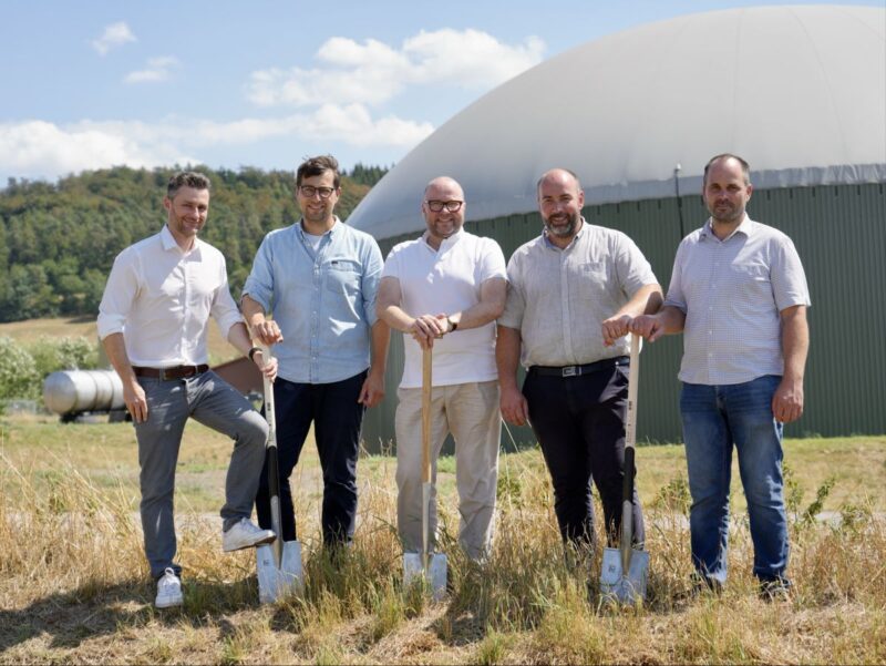 5 Personen mit Spaten vor einem Biogasbehälter.