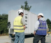 Zwei Personen stehen in Warnwesten und mit Baustellenhelm vor einer Speichereinheit.
