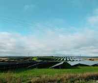 Blick über eine Freiflächensolaranlage auf grüner Wiese unter einem blau-bewölktem Himmel.