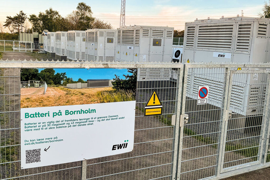 Großbatterieanlage mit Containern hinter einem Tor. Eine daran montierte Infotafel erklärt die Bedeutung der Anlage.