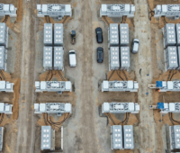 Luftaufnahme eines Großbatteriespeichers auf einer Baustelle. Mehrere Reihen von Batteriecontainern und technischen Umrichtereinheiten sind auf einem geschotterten Gelände angeordnet. Dazwischen stehen mehrere Fahrzeuge.