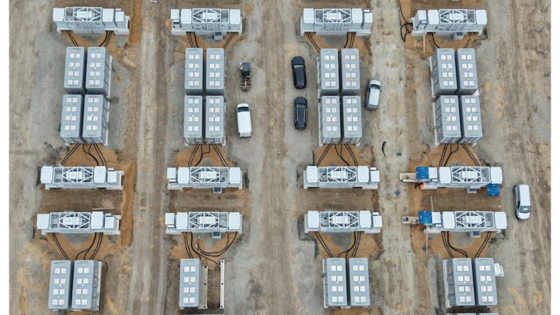 Luftaufnahme eines Großbatteriespeichers auf einer Baustelle. Mehrere Reihen von Batteriecontainern und technischen Umrichtereinheiten sind auf einem geschotterten Gelände angeordnet. Dazwischen stehen mehrere Fahrzeuge.