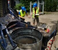 Nahaufnahme einer Brunnenbohrung auf einer Baustelle. Im Vordergrund ist das Bohrgerät mit einem Bohrrohr zu sehen, im Hintergrund stehen zwei Personen mit Schutzhelmen und Warnwesten auf dem Gelände des Pilotprojekts.