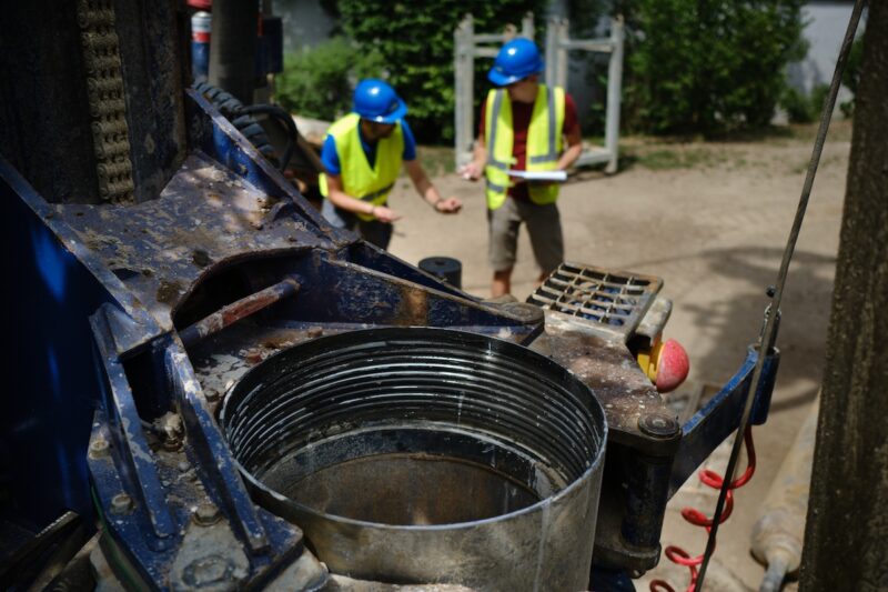 Nahaufnahme einer Brunnenbohrung auf einer Baustelle. Im Vordergrund ist das Bohrgerät mit einem Bohrrohr zu sehen, im Hintergrund stehen zwei Personen mit Schutzhelmen und Warnwesten auf dem Gelände des Pilotprojekts.