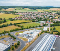 Luftaufnahme des H2-Hubs Hünfeld am Rand von Hünfeld-Michelsrombach. Die technische Anlage liegt zwischen einer Straße, Gewerbegebäuden und Feldern; im Hintergrund sind Wohnhäuser und eine bewaldete Hügellandschaft zu sehen.