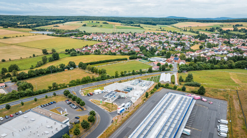 Luftaufnahme des H2-Hubs Hünfeld am Rand von Hünfeld-Michelsrombach. Die technische Anlage liegt zwischen einer Straße, Gewerbegebäuden und Feldern; im Hintergrund sind Wohnhäuser und eine bewaldete Hügellandschaft zu sehen.