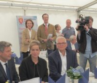 Drei Menschen (Habeck, Neubaur, Vogel) sitzen am Tisch in einem Zelt, im Hintergrund stehen zwei Menschen an der Zeltwand - Einweihung von Wald-Windpark