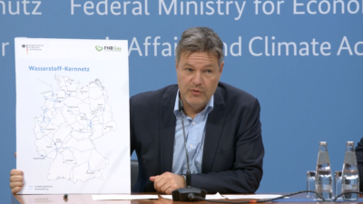 Mann mit Anzug - Robert Habeck - sitzt vor blauer Wand und zeigt Deutschland-Karte mit Linien - das Wasserstoff-Kernnetz.