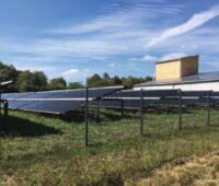 Hinter einem Zaun ist eine größere Solarthermieanlage mit Röhrenkollekoren installiert. Im Vordergrund eine grüne Wiese, im Hintergrund blauer Himmel. Am rechten Rang ragt ein kleiner Holzturm emport - er gehört zu einer Holzhackschnitzelanlage