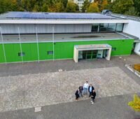 Luftbild mit fünf Personen vor einem öffentlichen Gebäude mit PV.