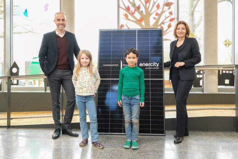 Zwei Kinder udn zwei Erwachsene vor einer Solaranlage in einer Schulaaula.