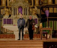 Das Foto zeigt zwei Personen vor dem Altar einer Kirche, die rechte Person hält eine Violine und Noten in der Hand.