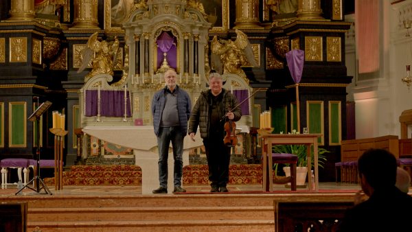 Das Foto zeigt zwei Personen vor dem Altar einer Kirche, die rechte Person hält eine Violine und Noten in der Hand.