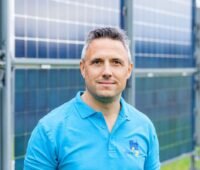 Mann im türkisfarbenen Polo-Shirt steht vor Vertikal-PV-Modulen, die unscharf im Hintergrund stehen.