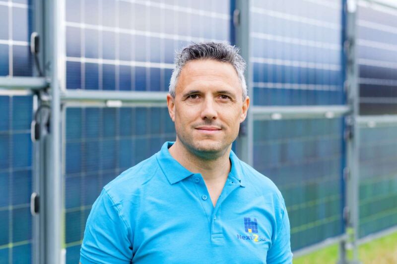 Mann im türkisfarbenen Polo-Shirt steht vor Vertikal-PV-Modulen, die unscharf im Hintergrund stehen.