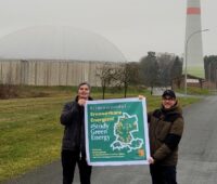 Zu sehen sind zwei Männer, die ein Plakat vom Netzwerk #StudyGreenEnergy tragen. Im Hintergrund eine Biogasanlage und ein Windkraftanlagenturm.