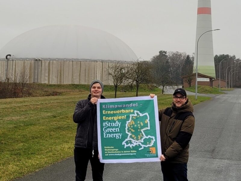 Zu sehen sind zwei Männer, die ein Plakat vom Netzwerk #StudyGreenEnergy tragen. Im Hintergrund eine Biogasanlage und ein Windkraftanlagenturm.