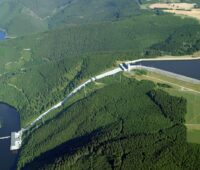Zusehen ist ein Pumpspeicherwerk aus der Vogelperspektive, welches einen höher gelegenen Stausee mit einem tiefer gelegenen Fluß verbindet.