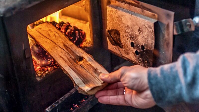 Eine Hand legt ein Holzscheit in einen Holzofen - Symbobild für Biomasse, Holzfeuerung, Feinstaub