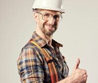 Mann mit weißem Bauhelm und großer Brille hält den Daumen hoch.