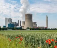 Großes Kohlekraftwerk mit mehreren Kühltürmen und Schornsteinen hinter einem Getreidefeld mit roten Mohnblumen. Aus einem Kühlturm steigt eine weiße Dampfwolke auf.