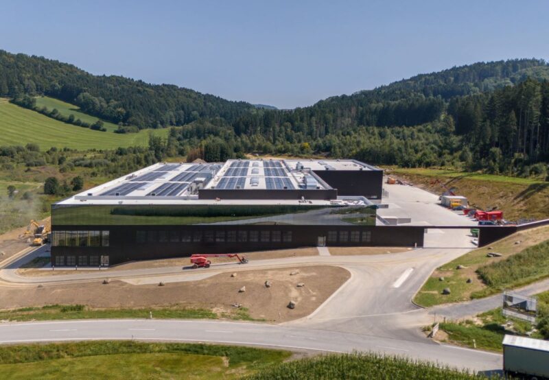 Neu gebautes Industriegebäude mit Photovoltaik auf dem Dach, umgeben von hügeliger grüner Landschaft.