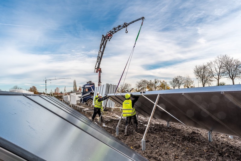 Monteure in gelben Westen installieren große Solarwärmeanlagen. Dabei hilft ein Kran, der im Hintergrund agiert.