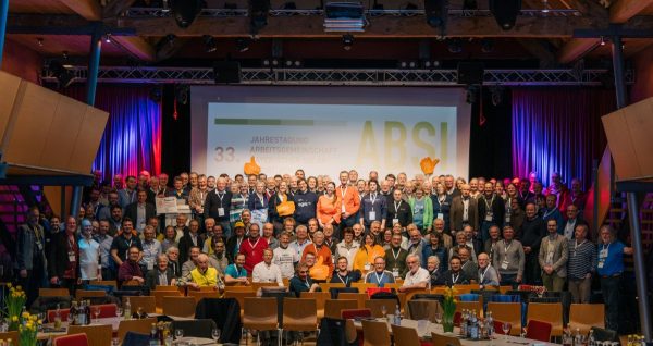 Das Foto zeigt eine Gruppenaufnahme in der Veranstaltungshalle vor der Bühne mit Projektion im Hintergrund.