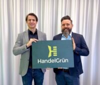 Die beiden Geschäftsführer halten ein dunkelgrünes Schild in der Hand mit einem hellgrünen H als Logo, darunter steht HandelGrün.