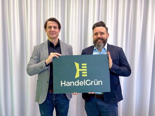 Die beiden Geschäftsführer halten ein dunkelgrünes Schild in der Hand mit einem hellgrünen H als Logo, darunter steht HandelGrün.