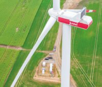 Drohnenfoto einer WIndkraftanlage mit Aufschrift "Juwi", im Hiuntergrund (unzu Füßen der Anlage im Hintergrund ein Container mit Batteriespeicher