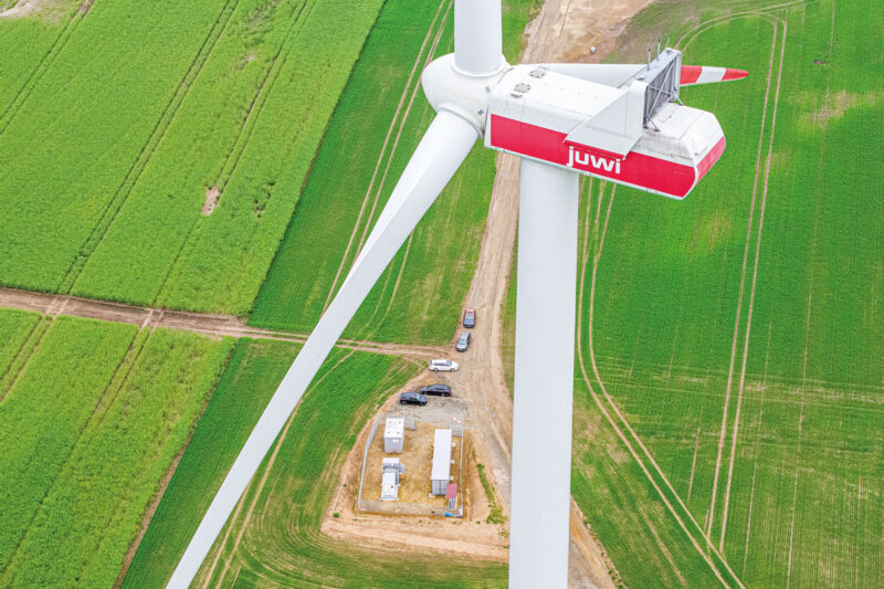 Drohnenfoto einer WIndkraftanlage mit Aufschrift "Juwi", im Hiuntergrund (unzu Füßen der Anlage im Hintergrund ein Container mit Batteriespeicher