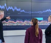 Grafische Benutzeroberfläche des WattPredictor an der Wand mit drei Personen davor: KI-gestützte Energieprognosen für Strom, Gas, Wärme und Kälte