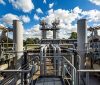 Eine Konstruktion aus grauen Pipelinen und Stangen mit zwei Männern in roter Weste im Hintergrund auf Türmen.