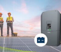 Grauer Batterie-Wechselrichter KOSTAL PLENTICORE BI 25 mit Batteriesymbol rechts vor zwei Monteuren mit gelben Warnwesten und orangen Schutzhelmen auf einer Photovoltaikanlage.
