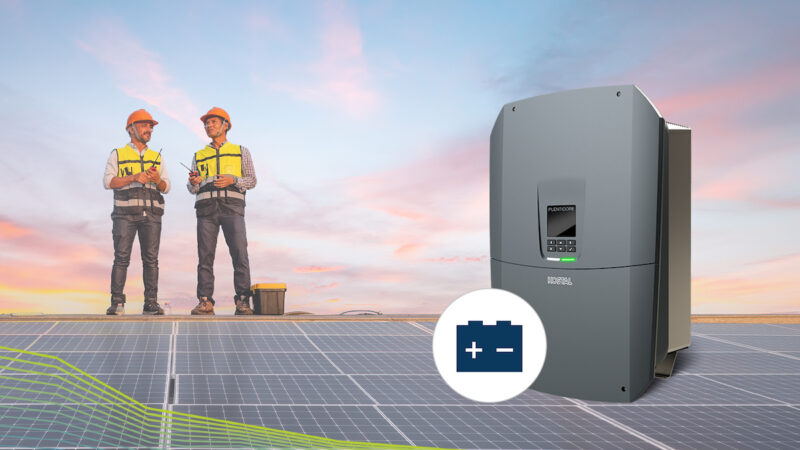 Grauer Batterie-Wechselrichter KOSTAL PLENTICORE BI 25 mit Batteriesymbol rechts vor zwei Monteuren mit gelben Warnwesten und orangen Schutzhelmen auf einer Photovoltaikanlage.