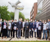 Ein Gruppe Absolventen (mit Maske) im Hintergrund das aufgeständerte Generatorhaus einer Windkraftanlage