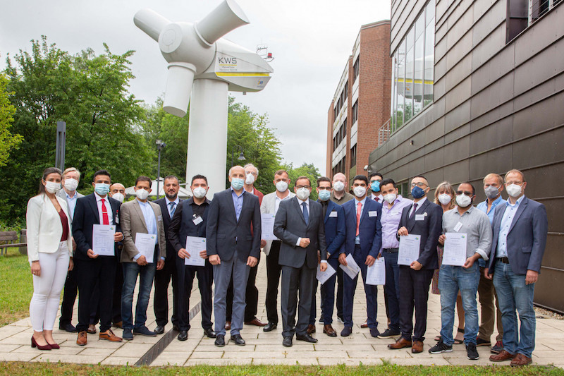 Ein Gruppe Absolventen (mit Maske) im Hintergrund das aufgeständerte Generatorhaus einer Windkraftanlage