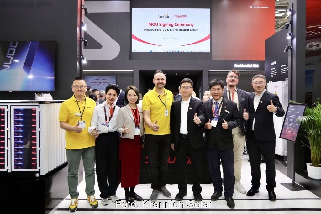 Eine Gruppe Menschen mit gelben T-Shirts und schwarzen Anzügen steht lachend vor einem Messestand.