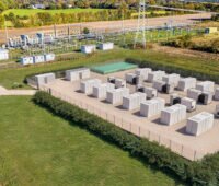 Luftaufnahme eines Batteriespeichers mit mehreren Container-Modulen auf einem eingezäunten Gelände neben einem Umspannwerk. Die Anlage ist von landwirtschaftlichen Flächen umgeben.