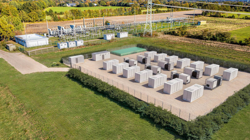 Luftaufnahme eines Batteriespeichers mit mehreren Container-Modulen auf einem eingezäunten Gelände neben einem Umspannwerk. Die Anlage ist von landwirtschaftlichen Flächen umgeben.