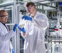 Im Bild zwei Männer im Labor mit einer Flasche des Energiespeichermaterials Preußisch Weiß für Natrium-Ionen-Batterien.